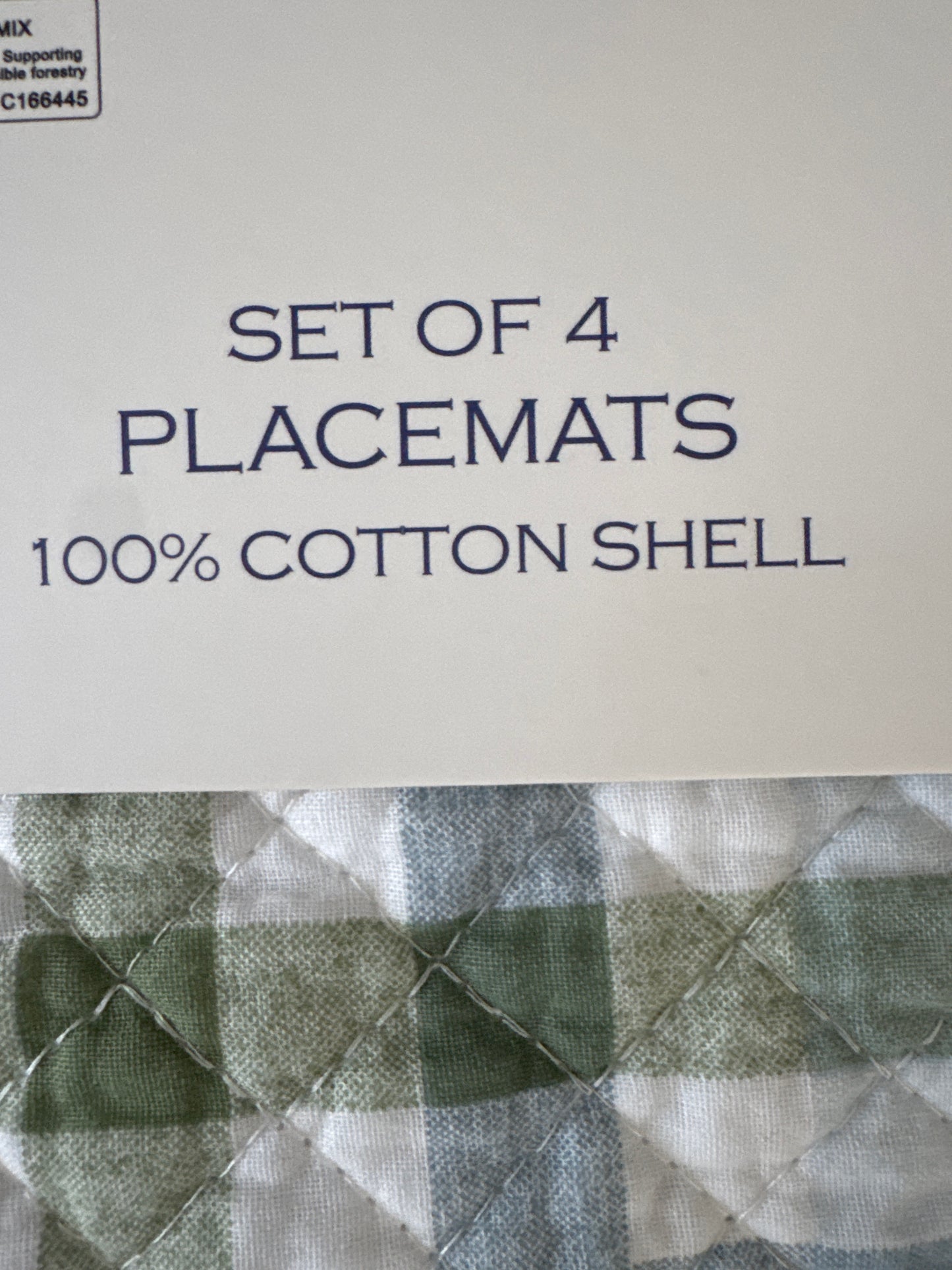 Close-up of the label on a set of 4 cotton placemats, displaying the text "SET OF 4 PLACEMATS 100% COTTON SHELL." The background shows a green and white plaid pattern on the placemats