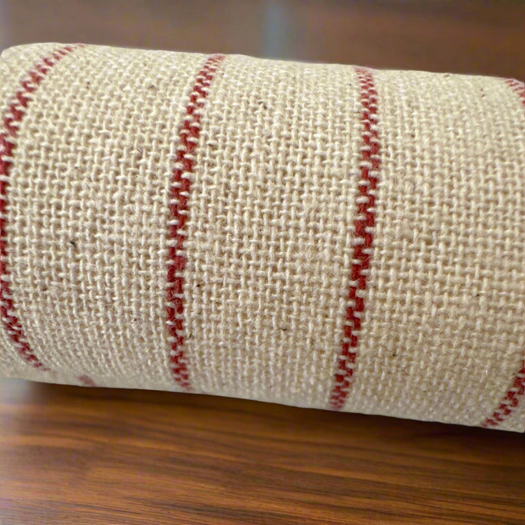 Rolled beige woven fabric with red parallel stripes, showcasing its textured surface and rustic appearance, resting on a wooden table